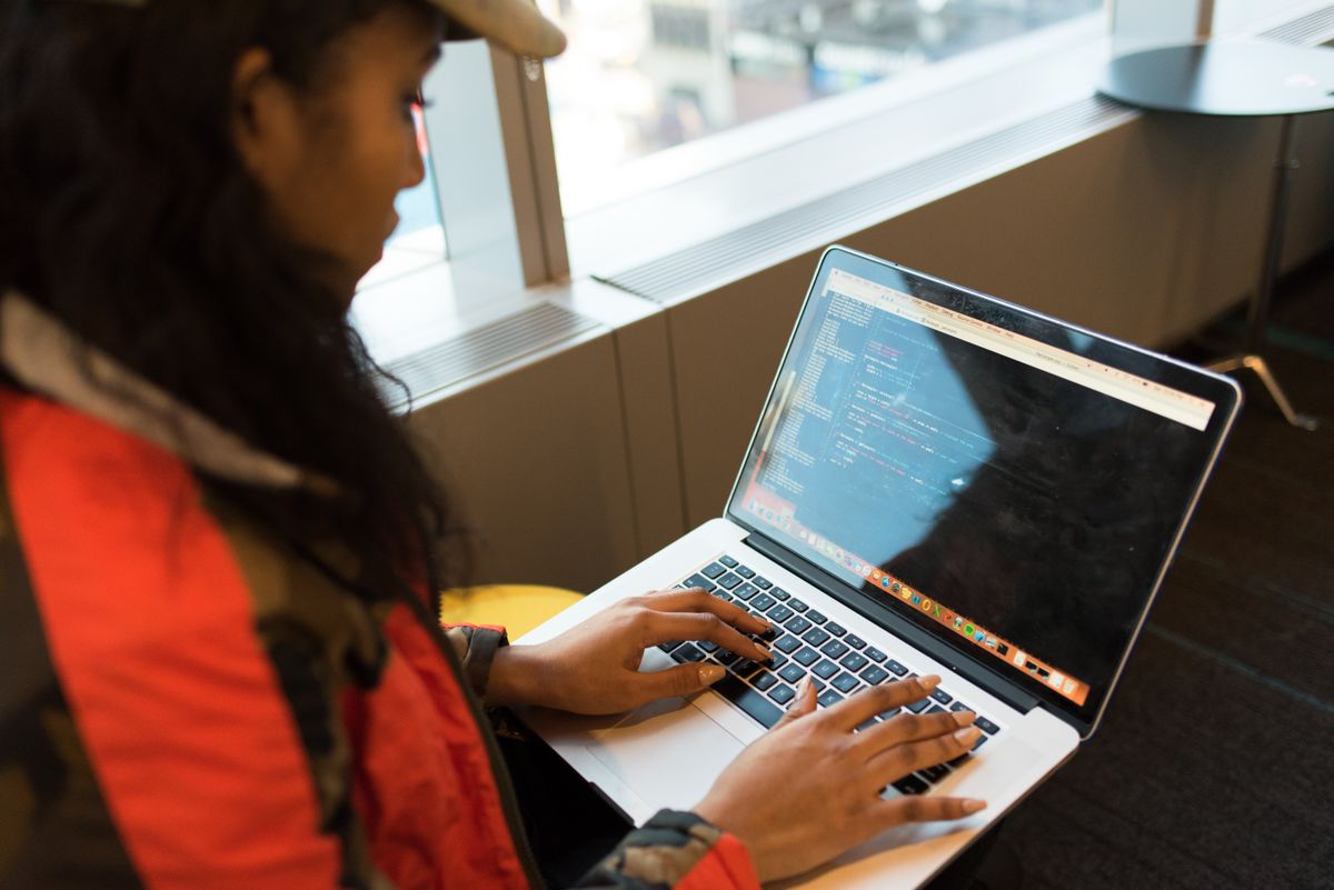 A young person writing code on a laptop beside a bright window.